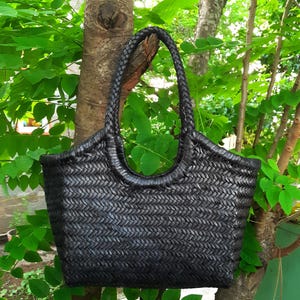 May include: A black woven tote bag with a braided handle. The bag is rectangular with rounded corners and a wide opening. The bag is hanging from a tree branch, surrounded by green leaves.