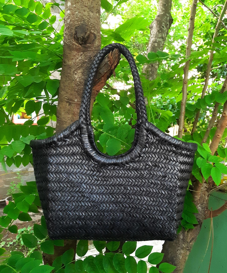 May include: A black woven tote bag with a braided handle. The bag is rectangular with rounded corners and a wide opening. The bag is hanging from a tree branch, surrounded by green leaves.