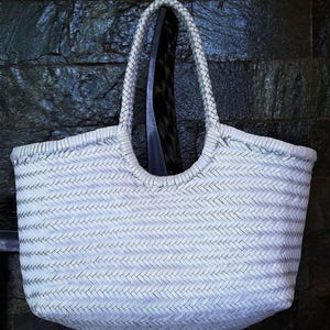May include: A white woven tote bag with braided handles. The bag has a rectangular shape with rounded corners and a large opening. The weave pattern is visible, and the bag appears to be made of a durable material. The bag is hanging against a dark background.