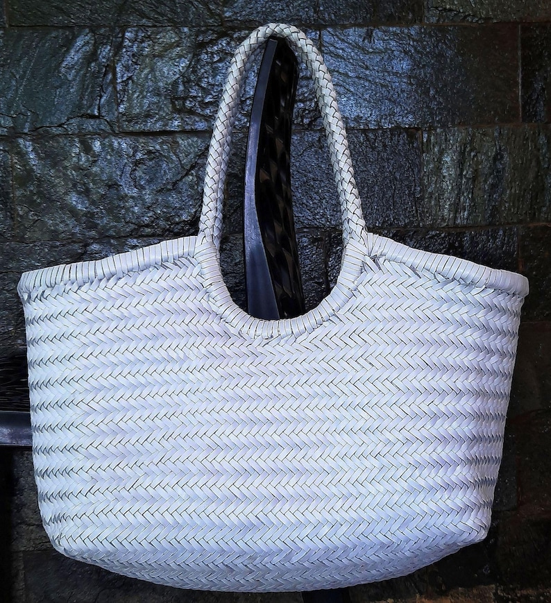 May include: A white woven tote bag with braided handles. The bag has a rectangular shape with rounded corners and a large opening. The weave pattern is visible, and the bag appears to be made of a durable material. The bag is hanging against a dark background.