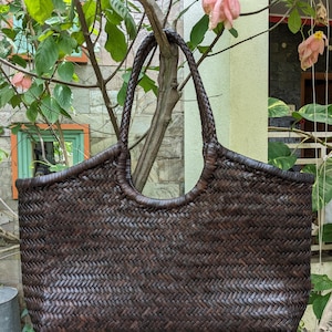 May include: A dark brown woven tote bag with a braided handle. The bag has a rectangular shape with rounded edges and a woven pattern. The bag is hanging from a tree branch, surrounded by green leaves and pink flowers.