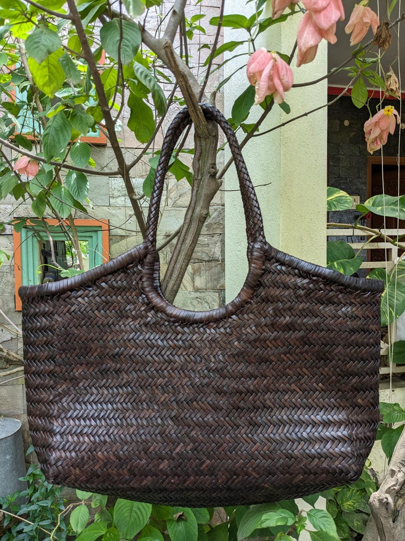 May include: A dark brown woven tote bag with a braided handle. The bag has a rectangular shape with rounded edges and a woven pattern. The bag is hanging from a tree branch, surrounded by green leaves and pink flowers.