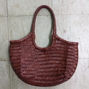 Puede incluir: Un bolso tote de color marrón oscuro que presenta un patrón de espiga trenzado en todo el cuerpo. El bolso tiene una forma de base redondeada y dos asas trenzadas integradas que se extienden desde la abertura superior, creando un aspecto uniforme.