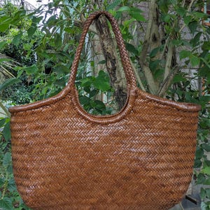 Peut inclure: Grand sac fourre-tout tissé marron avec une poignée tressée. Le sac a une forme arrondie et une surface texturée, avec un motif diagonal répétitif. Le sac est suspendu devant un fond de feuillage vert.