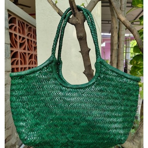May include: A woven green tote bag with braided handles. The bag has a rounded top and a textured, basket-weave design. The bag is hanging from a tree branch, showcasing its shape and color.