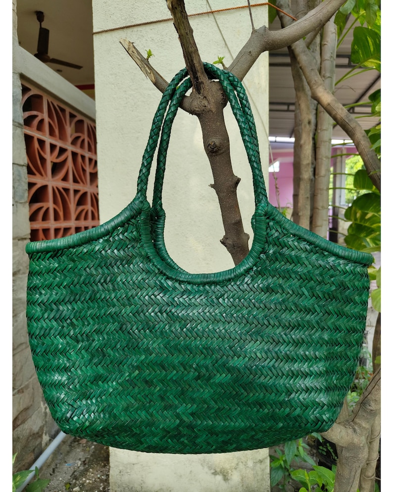 May include: A woven green tote bag with braided handles. The bag has a rounded top and a textured, basket-weave design. The bag is hanging from a tree branch, showcasing its shape and color.