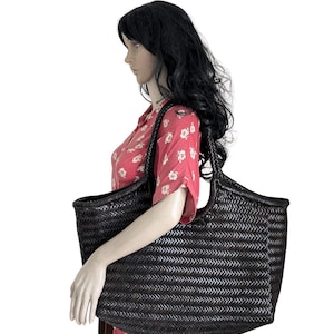May include: A large, dark brown woven tote bag with a wide opening and braided handles. The bag has a rectangular shape and a textured surface. The bag is held by a mannequin wearing a red floral dress.