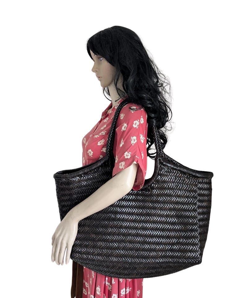 May include: A large, dark brown woven tote bag with a wide opening and braided handles. The bag has a rectangular shape and a textured surface. The bag is held by a mannequin wearing a red floral dress.