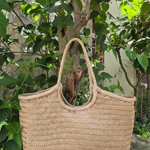 May include: A woven straw tote bag with braided handles. The bag has a rectangular shape with a wide opening and a natural beige color. The bag is set against a backdrop of green foliage and a tree trunk.