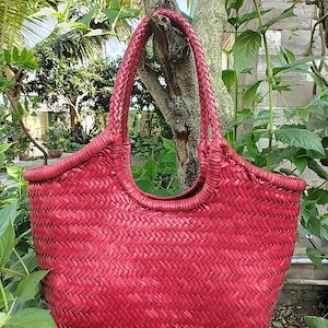 May include: A red woven tote bag with a braided handle. The bag has a rounded top and a textured, basket-weave design. The bag is hanging from a tree branch, surrounded by green foliage.