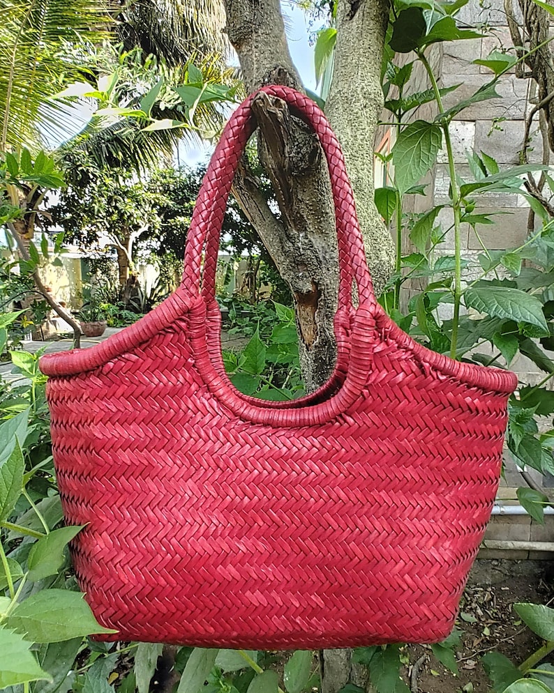 May include: A red woven tote bag with a braided handle. The bag has a rounded top and a textured, basket-weave design. The bag is hanging from a tree branch, surrounded by green foliage.