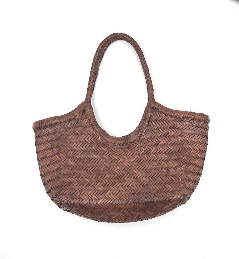 May include: A brown woven tote bag with a rounded base and a braided handle. The bag has a basket-like appearance, with a textured surface and a sturdy design. The handle is arched, and the bag is set against a white background.