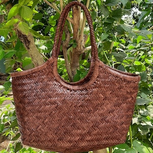 May include: A brown woven tote bag with braided handles. The bag has a wide, open top and a basket-weave pattern. The bag is hanging from a tree branch, with green leaves in the background. The bag is made of a durable material.