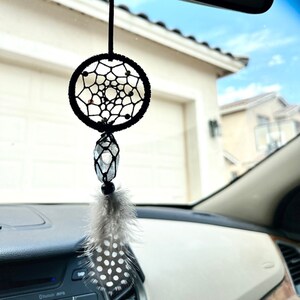 May include: Black dreamcatcher car charm with a crystal and a white feather with black polka dots hanging from the rearview mirror of a car.