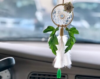 May include: A small dream catcher hanging ornament featuring a circular woven web, a beige fabric flower, two green serrated leaves, white feathers, and a single translucent green teardrop-shaped bead suspended from a thin brown cord.