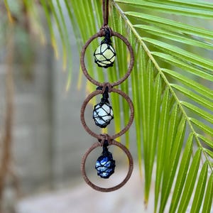 May include: A hanging decorative piece featuring three gemstone spheres encased in black netting, suspended within brown circular frames. The top sphere is a pale yellow, the middle is white, and the bottom is a deep blue. The piece is attached to a dark metal ring with a brown leather cord.