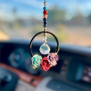 May include: A car charm featuring a black ring adorned with small fabric roses in teal, red, and purple. A faceted crystal hangs from the ring's center, and a strand of colorful beads tops the charm. The charm is hanging in front of a car dashboard.