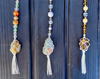May include: Three hanging ornaments featuring raw stones encased in white macrame netting with white tassels. Each ornament has a vertical strand of assorted beads in various colors, including green, orange, gold, clear, white, and brown tones.