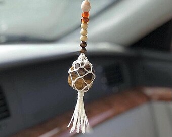 Puede incluir: Un adorno colgante para coche con una piedra ojo de tigre pulida en tonos marrón y dorado, envuelta en una red de macramé blanca. Sobre la piedra, una hilera vertical de cuentas en colores marrón oscuro, crema y ámbar termina en una borla.