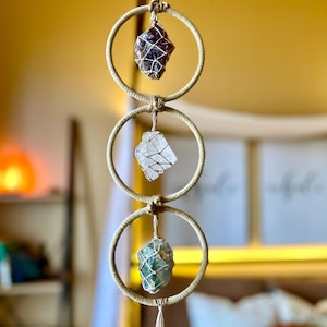 May include: A crystal mobile with three stones: amethyst, clear quartz, and green fluorite. Each stone is encased in a wire cage and suspended within a woven ring. The mobile hangs from a natural fibre cord, creating a bohemian aesthetic. The measurements are in centimetres.