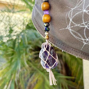 Puede incluir: Un colgante de cristal de amatista hecho a mano con una red de macramé, cuentas de madera y un anillo dorado. El colgante cuelga de un sombrero gris. La amatista es de color púrpura oscuro.