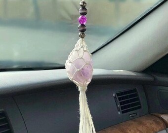 May include: A hanging car ornament featuring a light purple crystal encased in a white macrame net. The ornament is suspended by a string adorned with two dark grey beads, one bright pink bead, and silver-toned metal spacers, ending in a tassel.