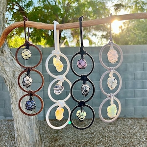 May include: Several crystal pendants, each with a different coloured gemstone held in a macrame net within a circular frame. The pendants, in shades of yellow, purple, pink, and white, are suspended from a wooden branch.