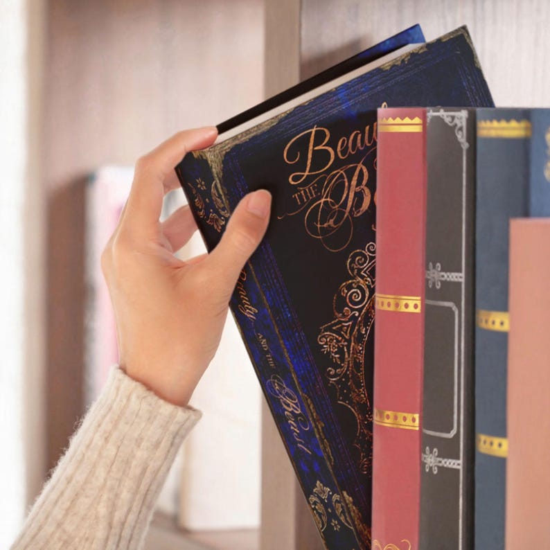May include: A hardback book titled Beauty and the Beast with a dark blue cover featuring gold filigree and ornate lettering, positioned on a shelf next to a red book with gold accents and a dark blue book with silver decorative patterns.