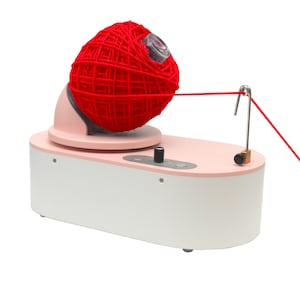 May include: A yarn ball winder machine with a large red yarn ball. The machine has a white and pink base with a silver metal arm and a black knob. The yarn is being wound from the ball.