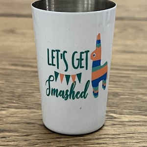 Puede incluir: Vaso de chupito blanco con borde plateado, con el texto "LET'S GET Smashed" en escritura verde y una colorida ilustración de piñata. La piñata tiene rayas naranjas, azules y verdes. Un artículo de fiesta divertido.