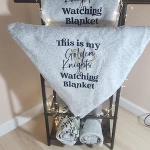 Puede incluir: Una manta de forro polar gris con el texto "This is my Golden Knights Watching Blanket" en un estante de metal. La manta está doblada y colocada sobre el estante. Dos mantas enrolladas están en el estante inferior.