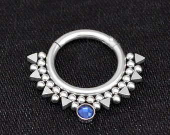 Puede incluir: Un anillo de joyería corporal de plata con una gema azul en el centro y una fila de cuentas y triángulos plateados alrededor del anillo.