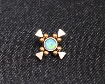 May include: A gold-toned body jewelry piece with a small, iridescent opal gemstone in the center. The piece is shaped like a cross with four triangular points and small round beads surrounding the gemstone.