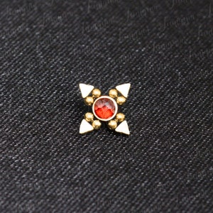 Può includere: Un gioiello per il corpo color oro con una pietra rossa al centro. Il pezzo ha quattro punte triangolari bianche che si estendono dal centro.