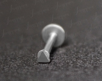 Pode incluir: Um piercing de nariz triangular prateado com uma parte de trás plana. O piercing está sobre um fundo preto.