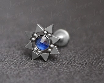 May include: A silver-toned stud earring with a starburst design. The earring features a central, round, deep blue gemstone surrounded by triangular and spherical accents. The earring post is visible.