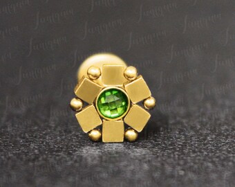 May include: Gold body jewelry with a green gemstone in the center. The jewelry is in the shape of a flower with eight petals.