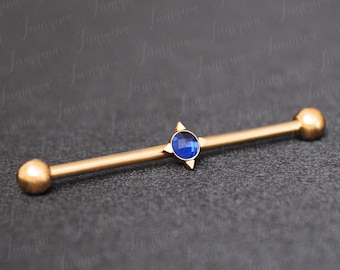 Puede incluir: Barbell de oro con una gema azul y un diseño en forma de estrella. El barbell es una joya de piercing corporal.