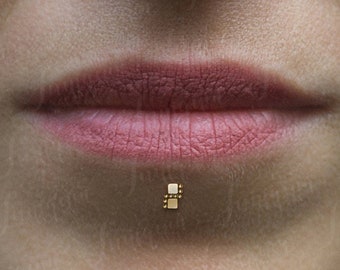 Puede incluir: Primer plano del rostro de una persona con un piercing labial dorado. El piercing tiene un diseño rectangular pequeño con un patrón de doble cuadrado. El tono de piel es claro y los labios son de color rosa natural.