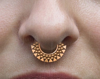 May include: Gold septum ring with a geometric design. The ring is made of metal and has a circular shape with a pointed edge.