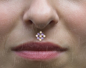 May include: Close-up of a person's face with a small, gold, flower-shaped nose piercing with purple gemstones.