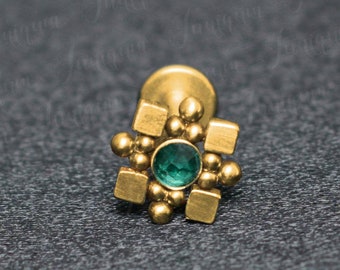 May include: Gold body jewelry with a green gemstone in the center. The jewelry is in the shape of a star with a square in the center.