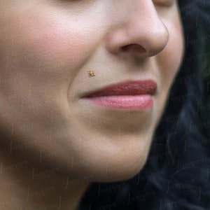 Op de afbeelding: Close-up van iemands gezicht met een kleine gouden oorbelpiercing aan de onderlip.