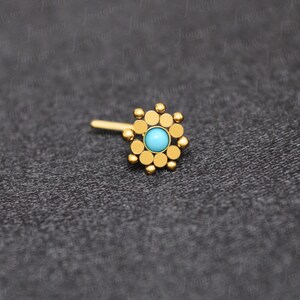 May include: Gold nose ring with a turquoise gemstone in the center and a circular design of small gold beads around the stone.