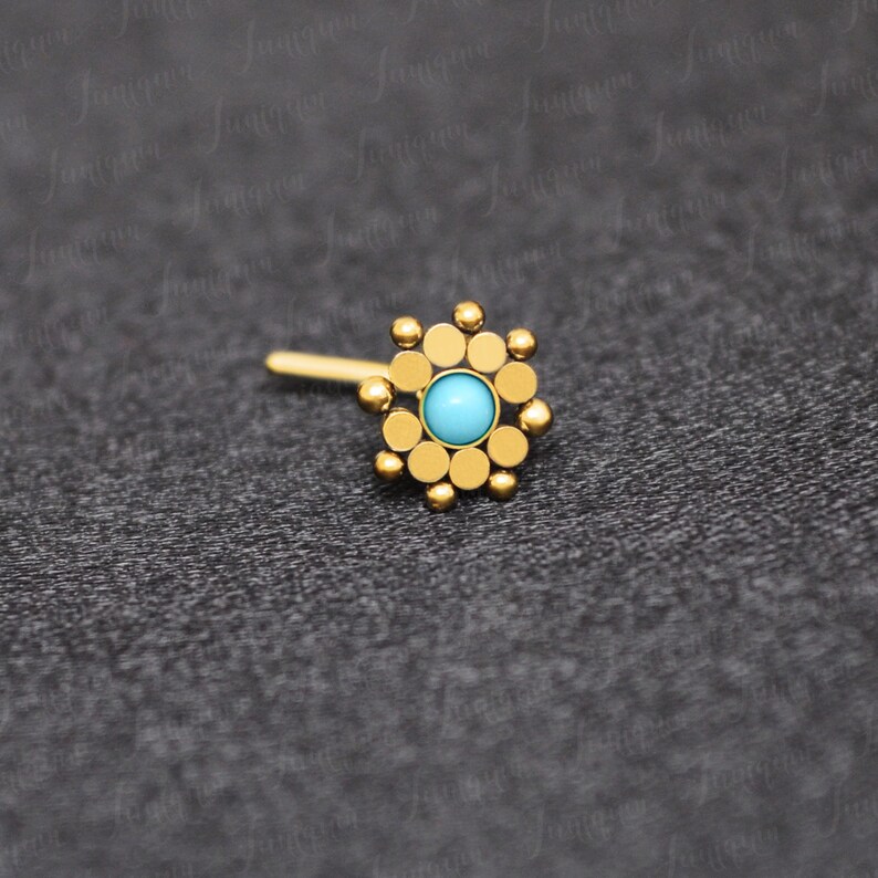 May include: Gold nose ring with a turquoise gemstone in the center and a circular design of small gold beads around the stone.
