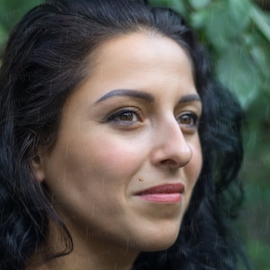 Op de afbeelding: Een close-up portret van een vrouw met lang, donker haar. Ze kijkt met een zachte glimlach naar de zijkant. Haar huid is licht en ze heeft bruine ogen. Ze draagt een kleine gouden oorbel in haar onderlip.