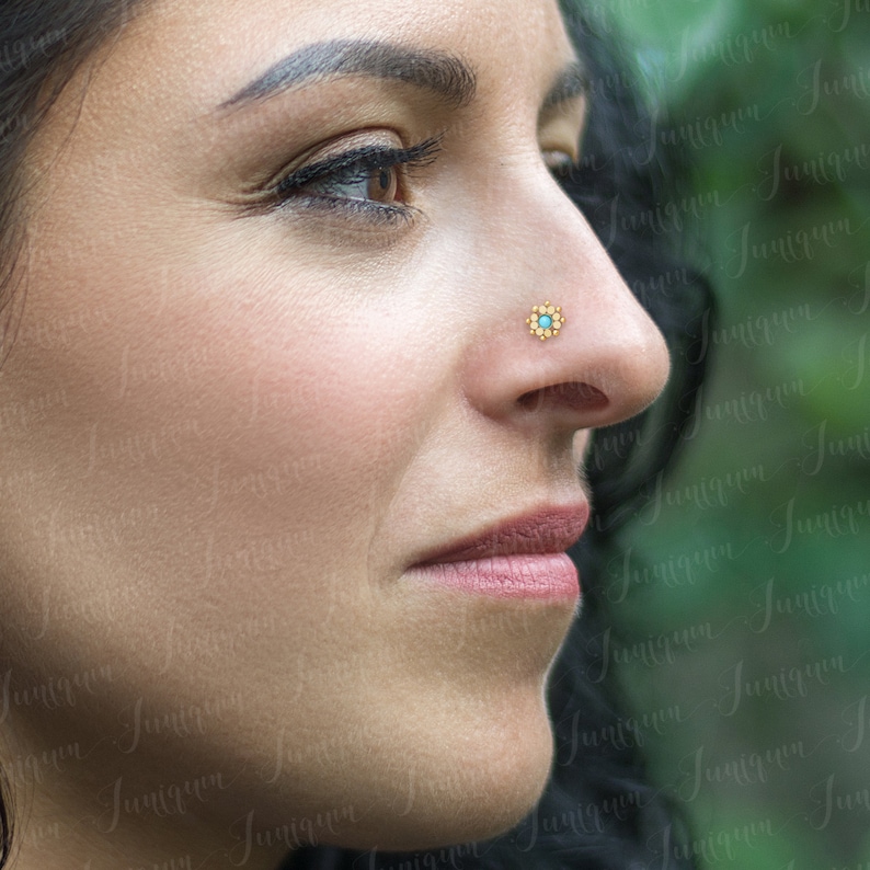 May include: A close-up of a person's face with a small gold nose ring featuring a turquoise gemstone in the center surrounded by a floral design.