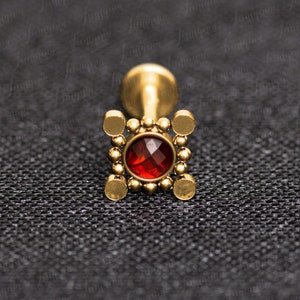 Pode incluir: Joia corporal de ouro com uma pedra preciosa vermelha engastada em uma moldura quadrada com pequenas contas douradas.