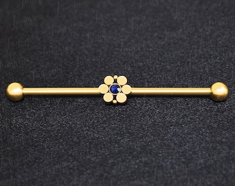 Puede incluir: Barbell de oro con un diseño de flor de gema azul. El barbell es una joya de piercing corporal.
