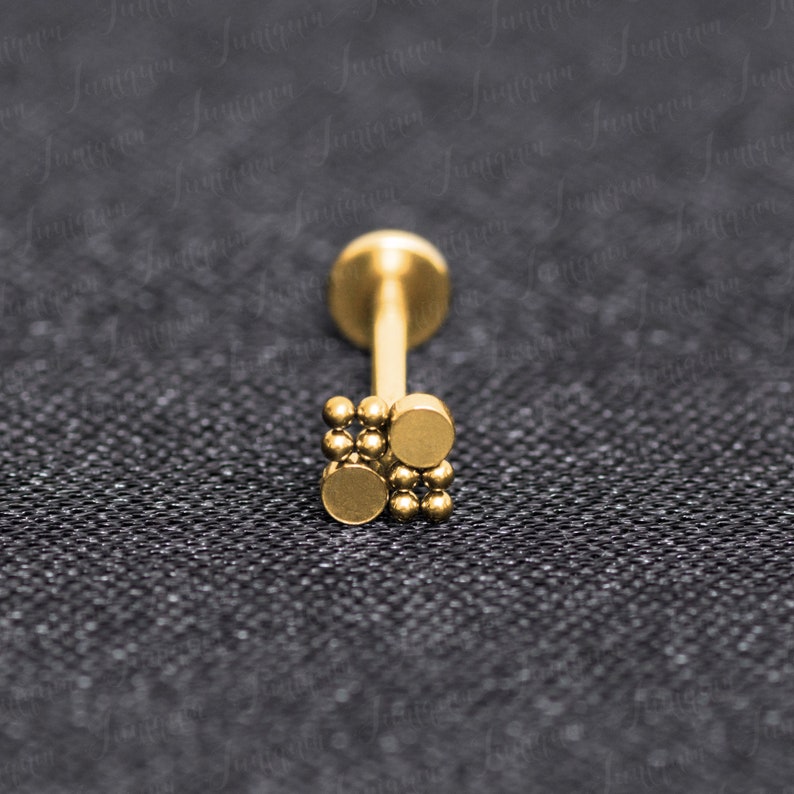 Op de afbeelding: Gouden lichaamsieraad met een klein, vierkant ontwerp met meerdere kleine balletjes. De sieraden zijn gemaakt van chirurgisch staal en geschikt voor verschillende piercings.